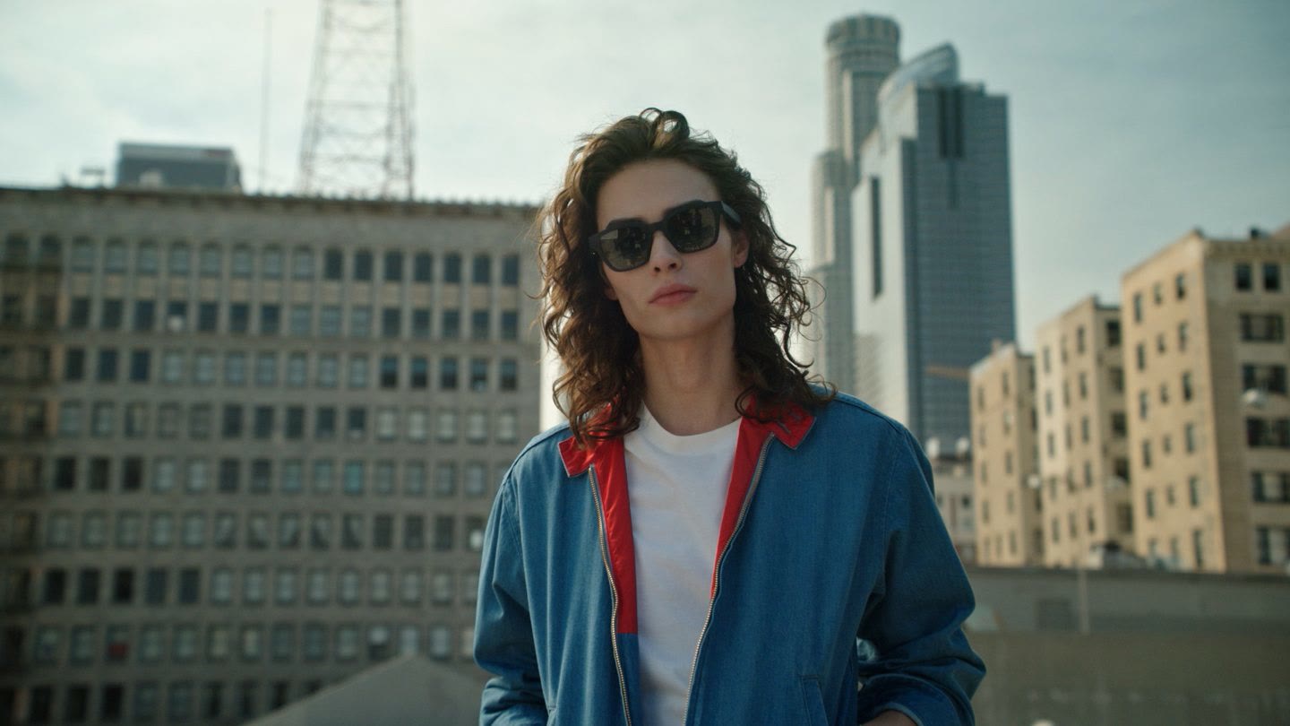 Rooftop portrait in daylight: a man with long curly hair and sunglasses, city towers behind.