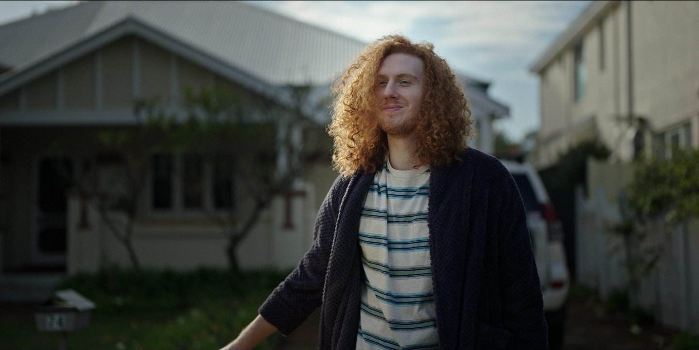Finished frame: a young man with curly red hair in a cardigan, standing in a suburban street in late sun, looking out of frame