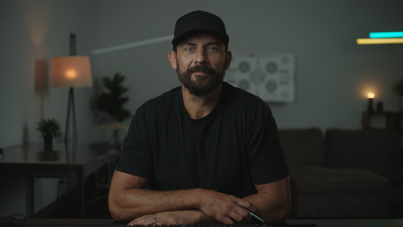 Patrick O'Sullivan seated at a desk in a dim room lit by a practical lamp, wearing a black cap and black T-shirt.