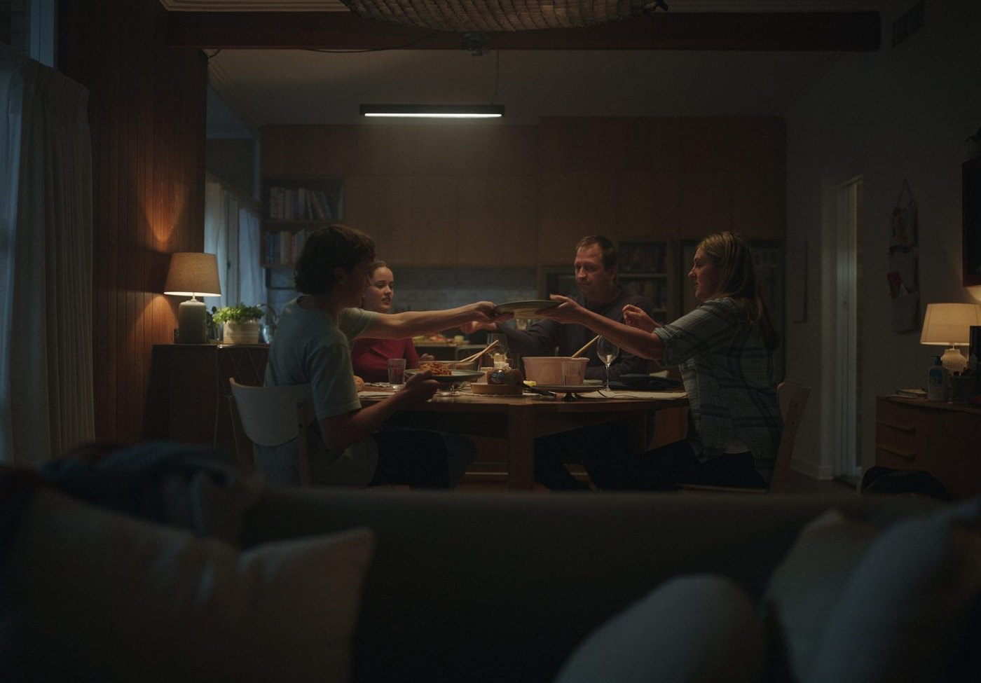 Finished frame: a family of four passing dishes around a dinner table at night, seen past a sofa in the foreground