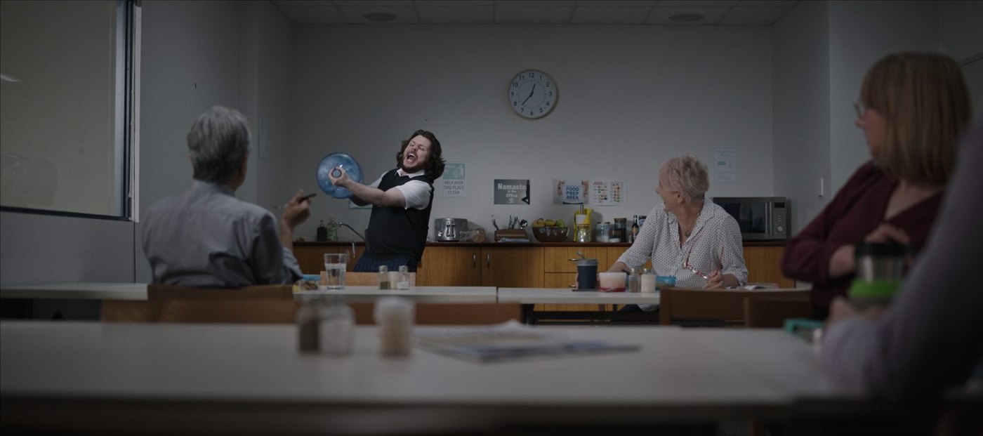 Finished wide frame: a man in a waistcoat celebrates with a water cooler bottle in an office lunch room while three coworkers at the tables watch