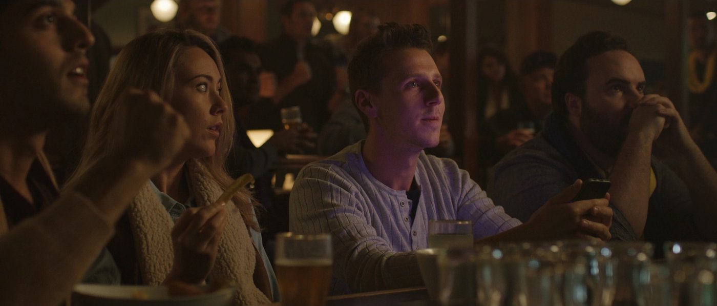 Finished frame: four people seated in a row at a pub bar, all facing the same way, watching something off screen