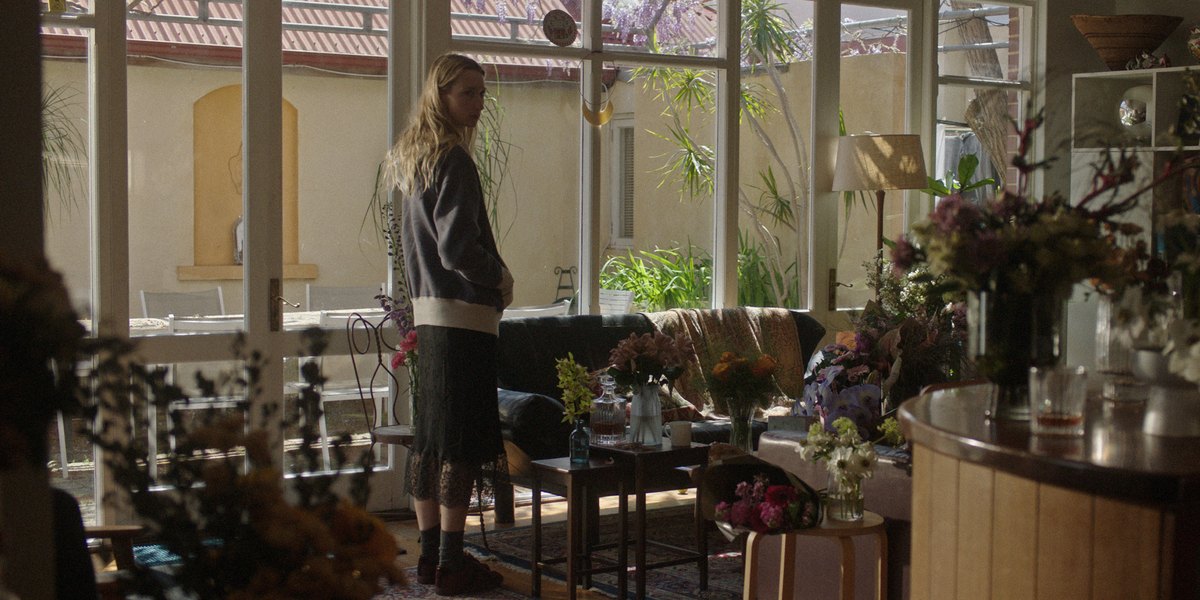 A woman standing in a sunroom full of flowers, light through the windows behind her